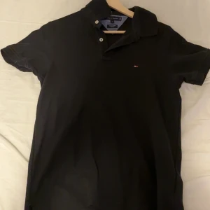 Svart pikétröja från Tommy Hilfiger - Klassisk svart pikétröja från Tommy Hilfiger med liten broderad logga på bröstet. Tröjan har korta ärmar, krage och knappar framtill. Perfekt för en clean och stilren look. Tillverkad i mjuk bomull som känns skön mot huden.