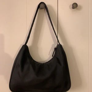 Svart axelväska från Monki - Snygg svart axelväska från Monki med enkel och stilren design. Väskan har en mjuk, lätt rundad form och dragkedja upptill. Insidan har ett extra fack med dragkedja för småsaker. Perfekt storlek för det viktigaste och passar till många outfits. Kan förekomma tecken på användning, men sparsamt använd.