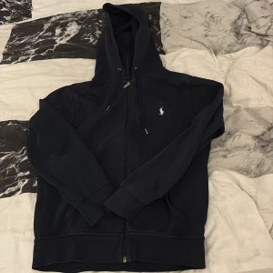Marinblå hoodie från Ralph Lauren - Snygg marinblå hoodie från Ralph Lauren med dragkedja och klassisk vit broderad logga på bröstet. Tröjan har huva med snörning, känguruficka och ribbade muddar. Säljer eftersom den inte längre används.