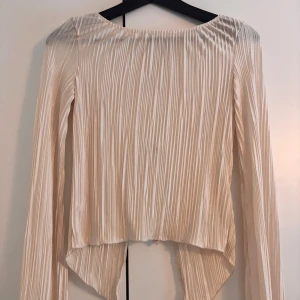 Beige blus från NA-KD - Säljer en elegant beige blus från NA-KD med långa ärmar och knytning baktill. Blusen har ett plisserat mönster och är tillverkad i ett glansigt material som ger en lyxig känsla. Perfekt för att höja din stil till nästa nivå!