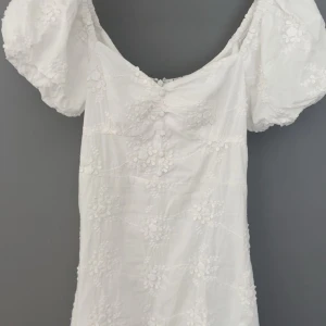 Vit kortklänning från Nelly - Supersöt vit kortklänning från Nelly med broderade blommor och puffärmar. Klänningen har dragkedja i ryggen och volangkant nertill. I storlek 36!