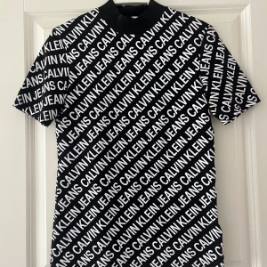 Svart t-shirt Calvin Klein Jeans XS - Svart t-shirt från Calvin Klein Jeans i storlek XS. T-shirten har korta ärmar och är täckt av ett diagonalt vitt logomönster över hela plagget. Klassisk passform och rund halsringning. Använd en gång 