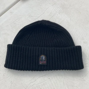 Svart ribbstickad mössa Parajumpers - Svart ribbstickad mössa från Parajumpers med uppvikt kant och klassisk PJS-logga framtill. Tillverkad i mjukt ullmaterial som håller dig varm. Enkel och stilren design som funkar till de flesta outfits.