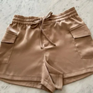 Beige shorts med snörning och fickor - Snygga beige shorts med elastisk midja och snörning framtill. De har två stora sidofickor med lock som ger en cool utility-vibe. Materialet känns mjukt och lite glansigt, perfekt för varma dagar. Passformen är relaxed och shortsen är lite längre i modellen.