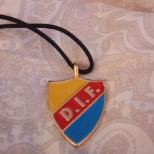 DIF halsband med klubbmärke - Halsband med Djurgårdens IF:s klassiska klubbmärke i sköldform. Märket har gul, röd och blå emalj med texten D.I.F. på röd bakgrund. Hängs i ett svart snöre och har guldfärgade kanter. Perfekt accessoar för dig som älskar Djurgården!