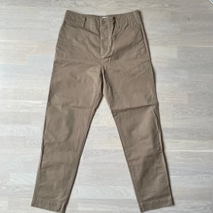 Beiga chinos från Filippa K, stl 46 - Snygga beiga chinos från Filippa K i klassisk rak modell. Byxorna har två sidfickor fram och två bakfickor med lock och knapp. Tillverkade i bomull med bälteshällor och knappgylf. Perfekta för en clean och stilren look.