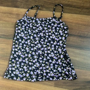 Blommig linnetopp från Lindex - Supersöt linnetopp från Lindex med smala axelband och svart botten täckt av lila och gula blommor. Perfekt för varma dagar och enkel att matcha med jeans eller shorts. Tunt och mjukt material som känns skönt mot huden.