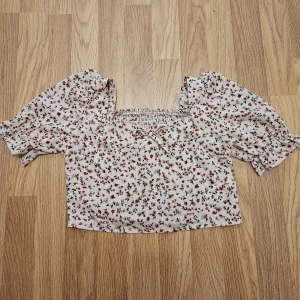 Blommig croppad blus med puffärm - Supersöt croppad blus i ljus färg med små röda blommor och gröna blad. Toppen har puffiga korta ärmar, smockad rygg och en liten rosett framtill. Perfekt för dig som gillar romantisk och trendig stil.