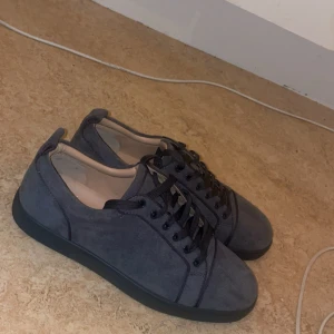 Grå Christian Louboutin  - Snygga sneakers i mörkgrå mocka från Christian Louboutin med klassisk röd sula. Skorna har svarta snören, rund tå och diskreta sömdetaljer. Perfekta för dig som vill ha en stilren men ändå unik look.