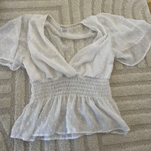 Vit blus med v-ringning och volang - Superfin vit blus med v-ringning och korta, luftiga ärmar. Blusen har små prickar i tyget, smockad midja och volang nertill. Perfekt för en fräsch och romantisk look till sommaren eller våren.