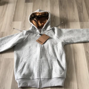 Grå hoodie från Burberry med dragkedja - (Stor leken är S men passar xs) Jag köpte den idag men den passade inte. Snygg grå hoodie från Burberry med klassiskt rutigt foder i huvan. Tröjan har dragkedja framtill, fickor och justerbar huva med snörning. Perfekt för dig som gillar stilrena och exklusiva plagg med en twist.