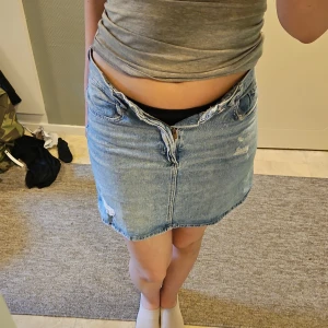 Blå jeanskjol med slitningar - Säljer en klassisk blå jeanskjol i kort modell med råa kanter och slitna detaljer framtill. Kjolen har fem fickor, bälteshällor och dragkedja med knapp. Perfekt för dig som gillar en avslappnad och trendig stil.