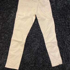 Vita jeans från J.Lindeberg Denim - Snygga vita jeans från J.Lindeberg Denim med klassisk femficksdesign och raka ben. Byxorna har hög midja, silverfärgade nitar och diskret logga bak på linningen. Perfekta för en clean och stilren look.