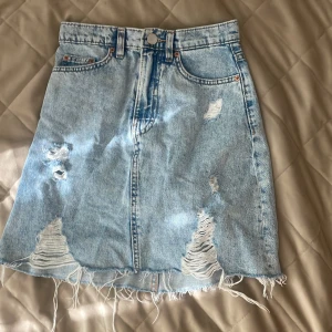 Ljusblå jeanskjol med slitningar - Säljer en ljusblå jeanskjol med rå kant och slitna detaljer framtill. Kjolen har klassiska fickor både fram och bak samt hällor för bälte. Perfekt för dig som gillar en avslappnad och trendig stil.