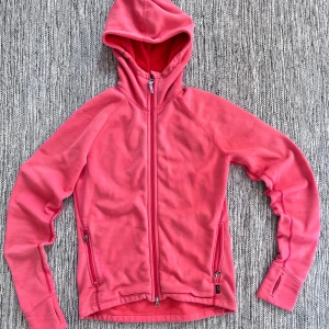 Houdini powerhoodie - Mysig rosa fleecejacka med huva och hel dragkedja framtill. Jackan har två fickor med dragkedja och tumhål vid ärmsluten. Perfekt för kyliga dagar när du vill vara både bekväm och snygg. Storlek S men passar lite större så dam m nästan 
