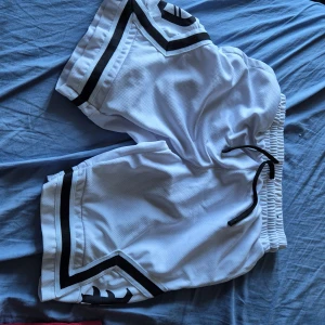 Vita sportiga shorts med svarta detaljer - Säljer ett par vita sportiga shorts med svarta ränder och coolt tryck på ena benet. De har elastisk midja med svart snörning och är gjorda i ett luftigt meshmaterial som passar perfekt för träning eller chill. Snygg kontrast mellan vitt och svart för en clean look.