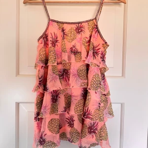 Rosa volanglinne med ananasmönster - Superfin ljus orange linne med volanger och tunna axelband. linnet har ett färgglatt ananasmönster och är perfekt för sommardagar. Lätt och luftig modell som ger en lekfull vibe.