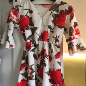 Vit blommig kortklänning med volangärm - Superfin vit kortklänning med stora röda blommor och gröna blad. Klänningen har v-ringning, omlottkänsla och volang på ärmarna. Perfekt för sommarens alla tillfällen och ger en romantisk vibe. Ingen lapp så osäker på storlek men skulle säga XXS