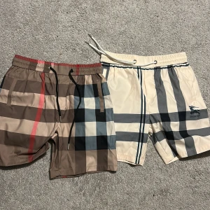 Burberry-rutiga badshorts med snörning - Två par klassiska Burberry badshorts med ikoniskt rutigt mönster. Ena paret är beige och brunt med röda detaljer, det andra är ljusbeige med grå och blå rutor samt Burberry-logga. Båda har elastisk midja och snörning för perfekt passform.