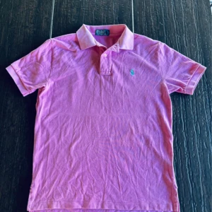 Rosa pikétröja från Polo Ralph Lauren - Snygg rosa pikétröja från Polo Ralph Lauren med klassisk krage och knappar framtill. Tröjan har korta ärmar och den ikoniska blå broderade loggan på bröstet. Perfekt för en chill och stilren look. Tillverkad i mjuk bomull som andas.