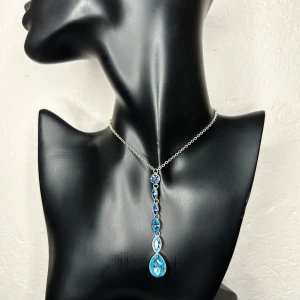 Y2K-halsband med blå kristaller – Glittrande 90-tal / 00-tals vintage - Ett snyggt halsband i silverfärg med flera blå gnistrande stenar i olika former, där den största är droppformad. Kedjan är tunn och elegant, och stenarna sitter på rad i ett coolt hängande mönster. Perfekt för dig som vill sticka ut med lite extra bling.