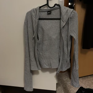 Grå ribbad hoodie från Gina Tricot - Grå ribbstickad hoodie från Gina Tricot i storlek XS. Tröjan har dragkedja framtill, huva med snörning och långa ärmar. Perfekt för en chill och stilren look, passar till både jeans och mjukisbyxor.