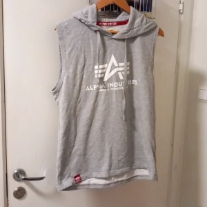 Grå ärmlös hoodie Alpha Industries - Snygg grå ärmlös hoodie från Alpha Industries med vit logga framtill och huva med snörning. Perfekt för lager-på-lager eller till gymmet. Mjuk bomullsblandning och relaxed fit ger en chill vibe.