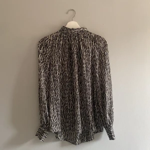 Zebramönstrad svartvit blus S - Snygg blus med zebramönster i svart och vitt. Blusen har lite högre krage med klädda knappar och långa ärmar med breda manschetter. Materialet är lätt och lite genomskinligt.