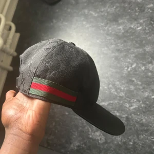 Svart Gucci keps med GG-mönster - Snygg svart keps från Gucci med diskret GG-monogram över hela kepsen och klassiska gröna och röda ränder på sidan. Kepsen har böjd skärm och är tillverkad i bomullsmaterial. Perfekt för dig som vill ha en lyxig och stilren accessoar.