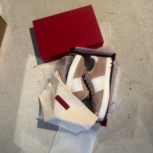 Flycrew Valentino Garavani beige sneakers mocka - Snygga sneakers från Valentino Garavani i beige mocka med vita skinnpaneler och vita snören. Sulan har coolt kamouflagemönster undertill och små nitar baktill. Kommer med originalkartong och dustbag. Perfekta för dig som gillar exklusiva detaljer och stilren design.