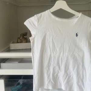 Vit t-shirt från Ralph Lauren - Klassisk vit t-shirt från Ralph Lauren med korta ärmar och rund hals. Ikoniska blå loggan broderad på bröstet. Skön och enkel modell i mjuk bomull, perfekt till jeans eller shorts.