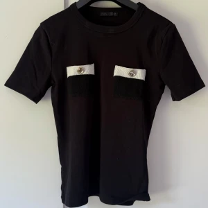 Svart t-shirt med kontrastfickor från Zara - Svart t-shirt från Zara med två coola bröstfickor i svart och vitt, dekorerade med silvriga knappar. T-shirten har rund hals och korta ärmar. Perfekt för dig som gillar en stilren men ändå unik look.