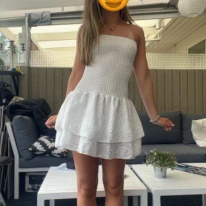 Vit axelbandslös kortklänning - Superfin vit axelbandslös klänning med smockad överdel och volangig kjol i två lager. Kjolen har broderade hålmönster som ger en söt och somrig känsla. Perfekt för varma dagar och midsommar! Säljes för den aldig har kommit till användning,Pris går att diskuteras