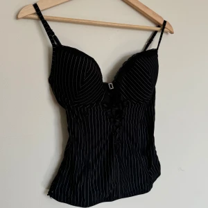 Svart randig korsett med snörning - Sart korsett med tunna vita ränder och smala axelband. Toppen har vadderade kupor, spetskant upptill och en liten glittrig detalj mellan kuporna. Framsidan har dekorativ snörning.