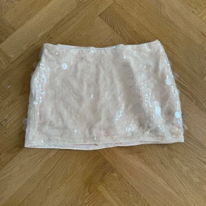 Beige paljettkjol i kort modell - Kort kjol i beige/vitt med glittrande paljetter. Kjolen har en rak passform. Materialet känns mjukt och följsamt, och paljetterna ger en riktigt festlig vibe.