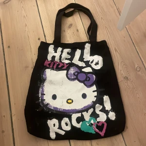 Hello Kitty shoppingväska med tryck - Svart shoppingväska från Hello Kitty i 100% bomull med stort tryck framtill. Motivet har vit, lila, rosa, turkos och gul färg och texten 'Hello Kitty Rocks!'. Väskan har långa handtag och är perfekt för att bära med sig grejer till skolan eller stan.