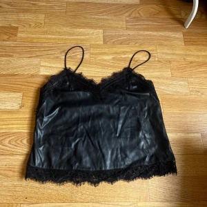 Svart linnetopp i skinnlook från Bik Bok - Snygg svart linnetopp från Bik Bok i skinnimitation med tunna axelband och spetsdetaljer längs v-ringningen och nederkanten. 🩷