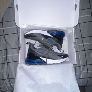 Nike Air Max 270 grå/blå sneakers - Nike Air Max 270 sneakers i grått med svarta och blå detaljer. Skorna har en stor blå Air Max-sula, mesh-ovandel och klassisk snörning. Perfekta för dig som gillar sportig stil och vill ha bekväma sneakers med modern design.