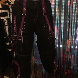 Svarta Tripp NYC cargopants med kedjor - Svarta cargopants från Tripp NYC med lila detaljer, nitar, kedjor och remmar. Byxorna har flera dragkedjor och fickor, samt coola metalldetaljer och lila sömmar. Perfekta för dig som gillar alternativ stil och vill sticka ut.