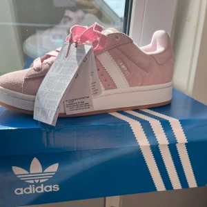 Adidas Campus 00s rosa sneakers - Snygga Adidas Campus 00s sneakers i ljusrosa mocka med klassiska vita ränder och vit sula. Skorna har rund tå, platt gummisula och rosa snörning. Perfekta för dig som vill ha en retro vibe med modern touch.