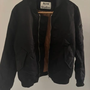Svart bomberjacka från Acne Studios - Svart bomberjacka från Acne Studios med klassisk siluett, orangefärgat innerfoder och grov dragkedja. Jackan har fickor med lock, ärmficka med dragkedja och ribbade muddar. Perfekt för dig som vill ha en stilren och tidlös look.