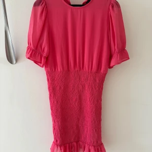 Rosa kort klänning - Superfin rosa kortklänning från Vero Moda med puffärmar, smockad kjol och volang nertill. Klänningen har smockad kjol och är gjord i ett lätt, syntetiskt material. Perfekt för dig som gillar färg. Rund halsringning och kort ärm. 