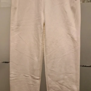 Beige mjukisbyxor från Champion S - Snygga beige mjukisbyxor från Champion i ekologisk bomull och återvunnen polyester. Byxorna har resår i benslut och broderad logga vid benet. Perfekta för chill eller träning, supermjuk känsla och relaxed fit.