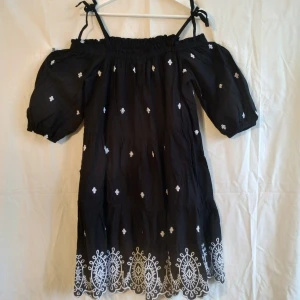 Svart kortklänning med broderi H&M - Supersöt svart kortklänning från H&M med vita broderade detaljer och puffärmar. Klänningen har offshoulder-modell med knytbara axelband och volangkant nertill. Perfekt för sommarens alla tillfällen!