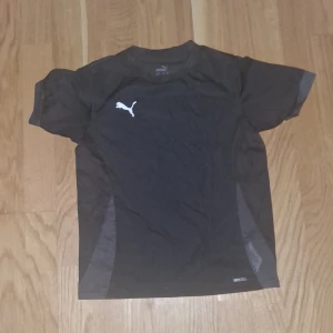 Svart sportig t-shirt från Puma - Svart t-shirt från Puma med vitt logotryck på bröstet. Tillverkad i ett lätt och ventilerande syntetmaterial, perfekt för träning. Klassisk passform med korta ärmar och diskreta meshpaneler på sidorna för extra andningsförmåga. 
