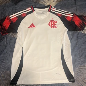 Vit Flamengo fotbollströja Adidas - Snygg vit Flamengo fotbollströja från Adidas med röda och svarta detaljer på ärmarna och klubbmärke broderat på bröstet. Tröjan har klassiska Adidas-ränder på axlarna och är tillverkad i lätt, ventilerande material (Aeroready). Perfekt för dig som älskar brasiliansk fotboll.