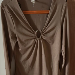 Brun långärmad topp med ringdetalj - Säljer en brun långärmad topp med djup v-ringning och snygg ringdetalj framtill. Toppen har en tight passform och är tillverkad i mjukt stretchigt material. Perfekt för dig som gillar trendiga detaljer och vill sticka ut lite extra.
