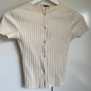 Beige ribbad topp med knappar - Snygg beige ribbad topp från Gina Tricot med korta ärmar och knappar hela vägen framtill. Toppen har en figurnära passform och är gjord i mjuk bomullsmix. Oanvänd utan prislapp.