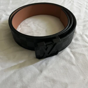 Louis Vuitton svart skärp med LV-spänne - Snyggt svart bälte från Louis Vuitton med klassiskt rutigt mönster och mattsvart LV-spänne. Bältet är gjort i exklusivt skinn och har en brun insida. Perfekt accessoar för att lyfta din outfit med en lyxig touch.