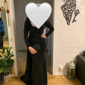 Svart glittrig långklänning - Elegant svart långklänning med glittrigt tyg och omlott-detalj. Klänningen har långa ärmar och markerad midja, vilket ger en snygg siluett. Perfekt för dig som vill sticka ut på festen. Skriv för mer bilder💞